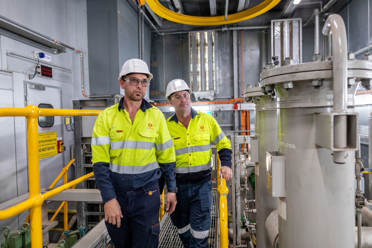 Two Alinta Energy workers in yellow hi-vis and hard hats proceed down a railed walkway onsite.