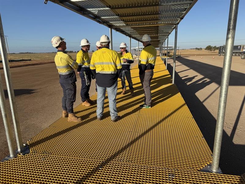 A group of workers in hi-vis clothing and hard hats standing on a yellow grated walkway under a metal canopy at an outdoor worksite, talking together in the sun.
