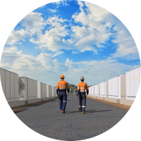 A CG image of two workers walking a path between two rows of large batteries. Above is a blue, cloudy sky.