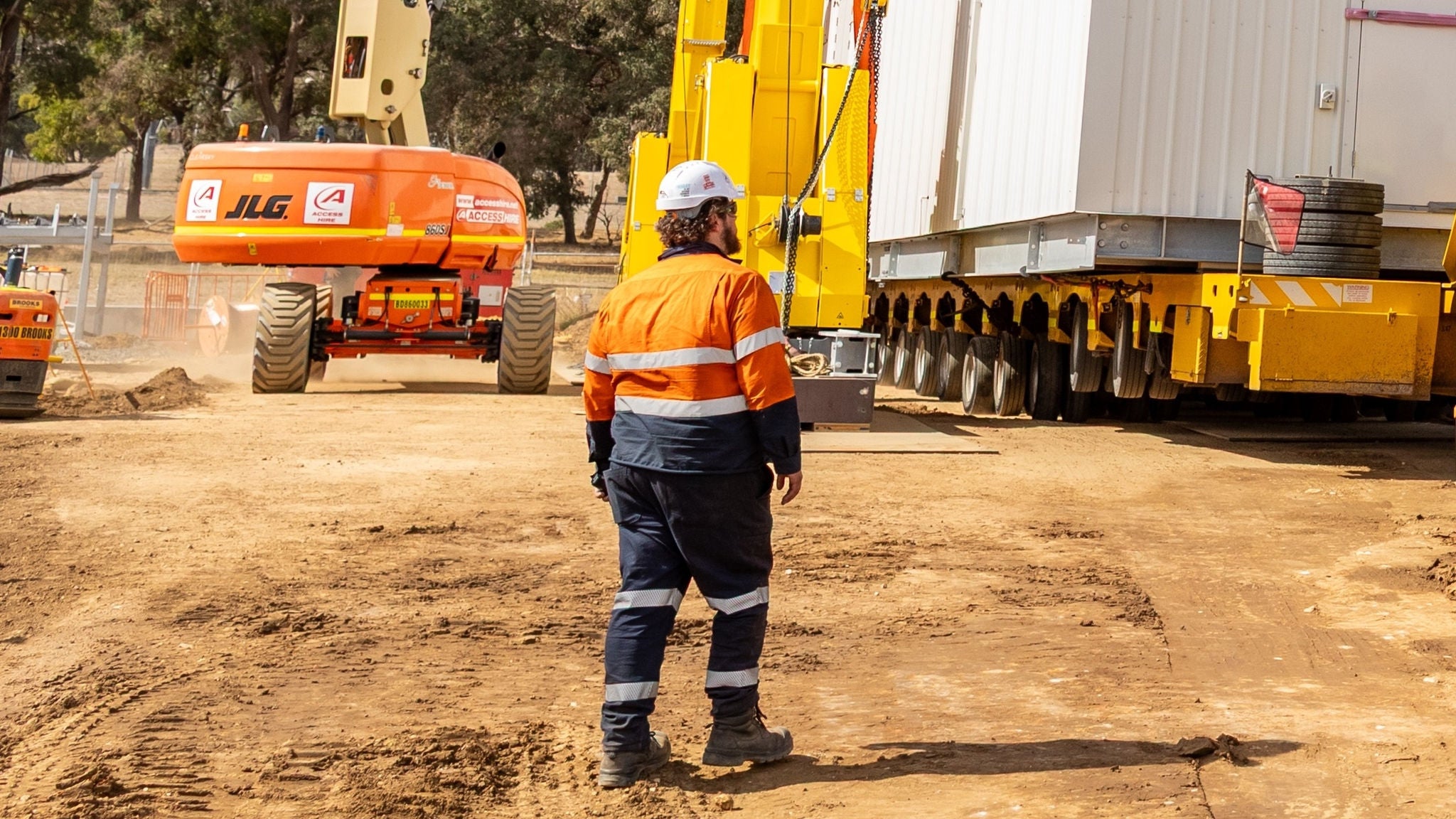 A worker in orange hi-vis and hard hat standing in front of construction machinery. He is seen from the back.