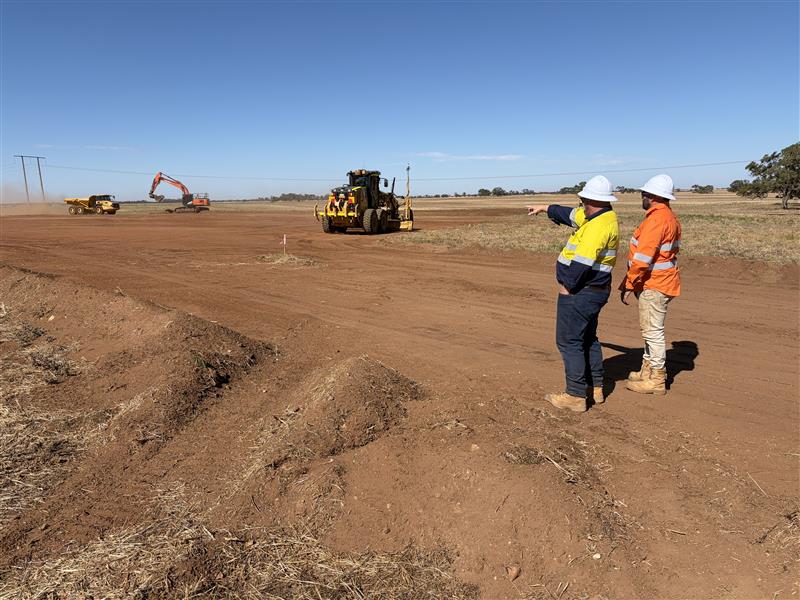 Two workers on a desert road. One wears yellow hi-vis and points to the horizon. The other wears orange hi-vis.